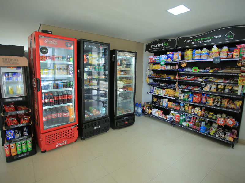 Uma gôndola com produtos usuais do dia a dia, ao lado de três geladeiras com bebidas, todas alocadas em uma franquia de minimercado