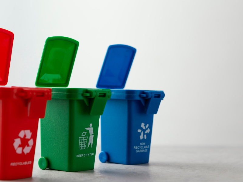 Três lixeiras lado a lado, a primeira na cor vermelha, a segunda na cor verde e a terceira na cor azul, exemplificando a separação do lixo nos condomínios