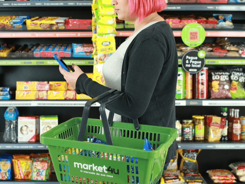 Mulher branco com o cabelo curto rosa em frente a uma gôndola de mercado autônomo do market4u segurando uma cesta de compras e um celular