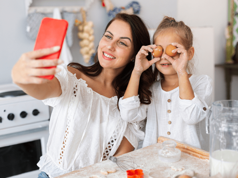 Mulher ao lado da filha tira uma selfie com o celular na mão enquanto a filha segura um ovo em cada mão tapando o os olhos