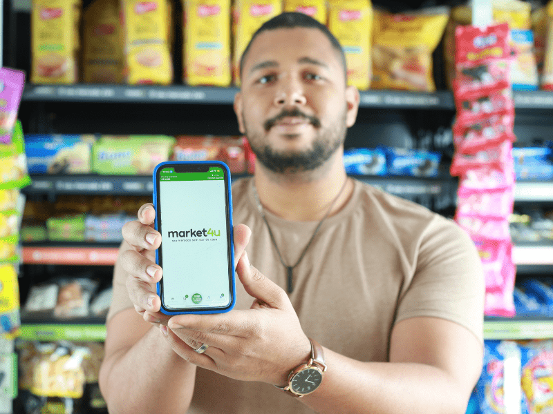 Homem negro segura um celular na mão, com a tela virada para frente. Na tela aparece o aplicativo do market4u, que está entre as melhores franquias de 2023. Ao fundo, uma gôndola do market4u