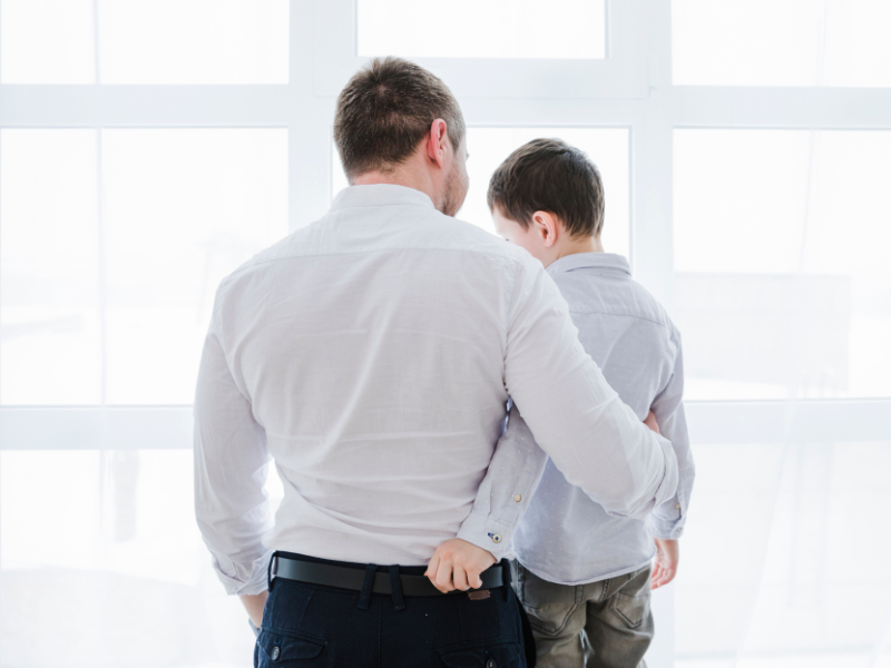 Homem branco de calça social preta e camisa clara, ao lado do filho branco, de calça jeans e camisa social clara. Ambos de costas enquanto o pai abraça o filho.