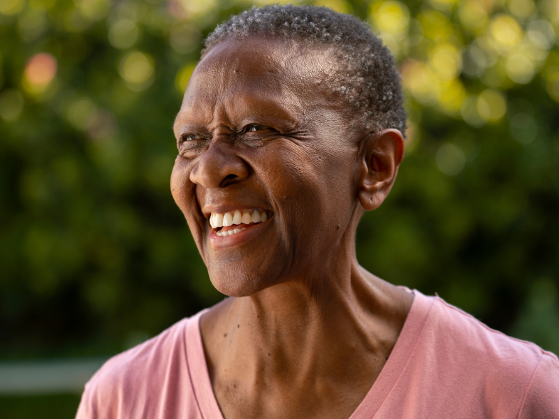 Mulher, negra, sorrindo em frente umas árvores, representando uma população mais idosa